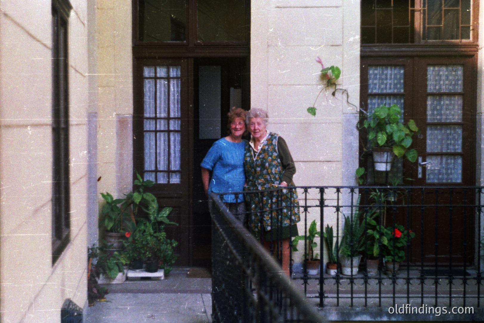 Two women stand on a balcony overlooking a traditional facade. Lush potted plants decorate the wrought-iron railing. The architectural style suggests a European location, possibly Mediterranean. Likely taken in the 1970s or 1980s. Offers a nostalgic glimpse of everyday life.