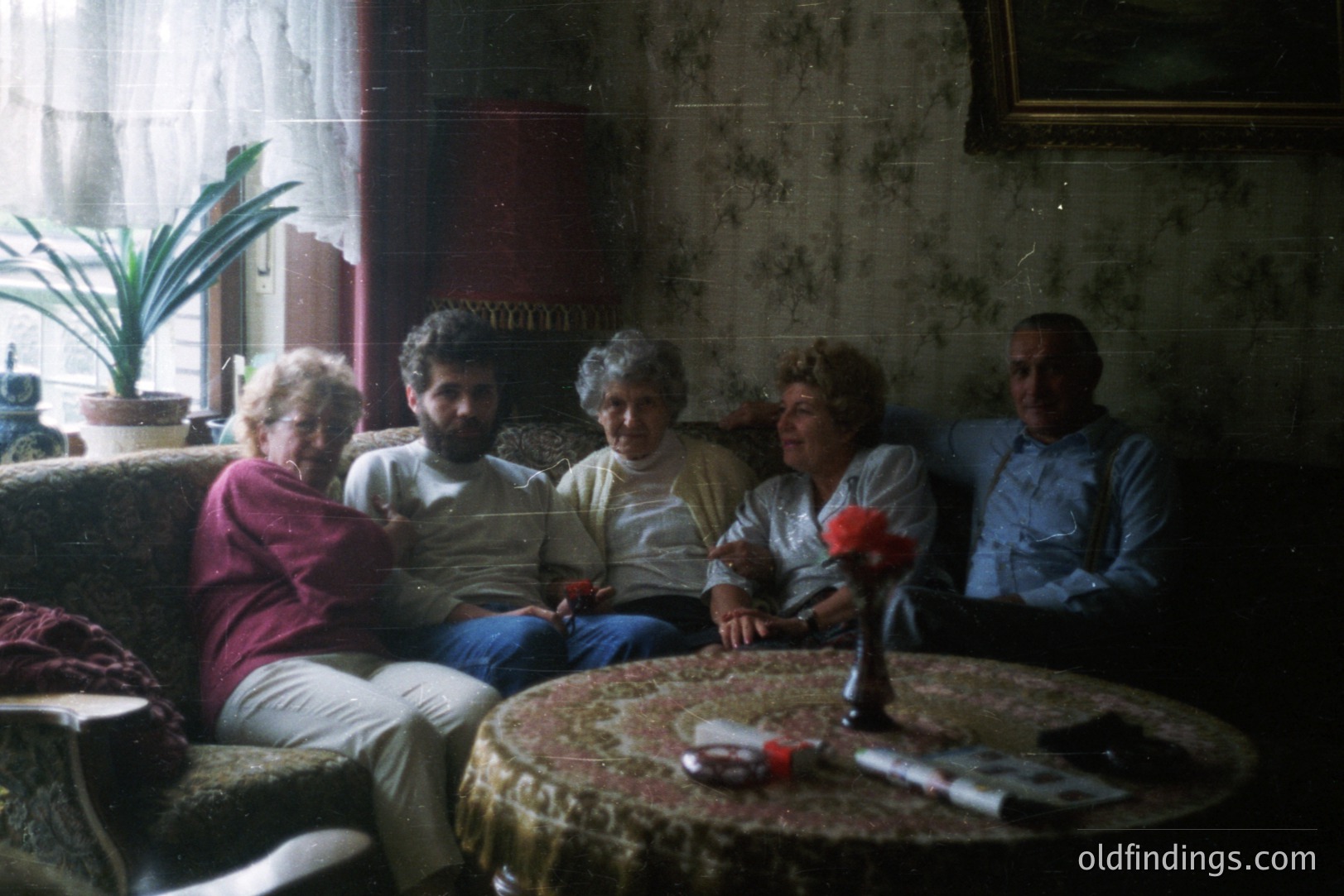 Four individuals seated on a floral-upholstered sofa in a vintage interior. A round table covered with an ornate cloth sits center. Visible details suggest a likely 1970s/80s era, possibly Eastern European domestic scene. Floral wallpaper and a potted palm add to the atmosphere.