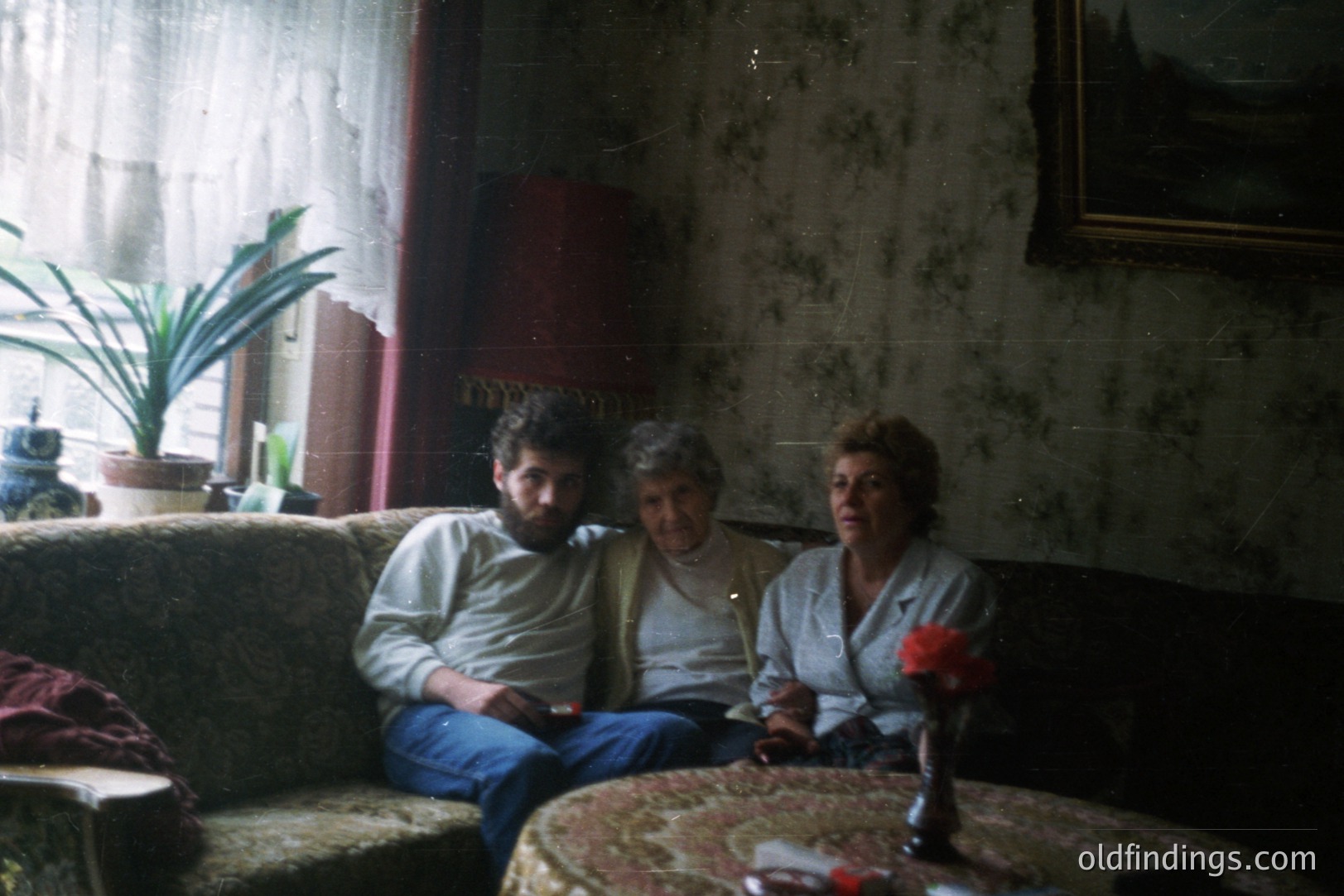 A family scene: a bearded man, older woman, and a younger woman sit on an ornate, floral-patterned sofa in a dimly lit room. Visible wall texture and a large, framed painting suggest a historic interior. Likely 1970s, Eastern European setting. The room’s decor indicates potential value for period design reference.
