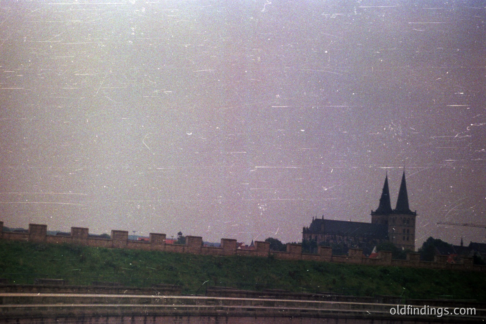 A view of a medieval castle with prominent twin towers, set atop a grassy hill. The castle is backed by a stone wall section. Heavily scratched film suggests age and potential archival value. Location & date unknown. Likely 1970s or 80s film stock.
