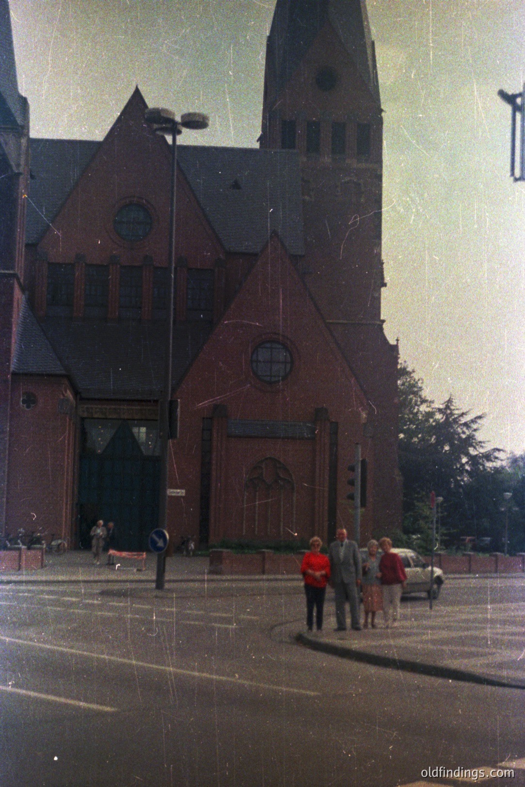 A striking architectural view of a brick church with a tall, pointed spire. Three formally dressed figures – a man in a suit, and two women – stand near a vintage automobile. Likely 1960s or 1970s, judging by the clothing and car style. The image shows weathering, suggesting age.