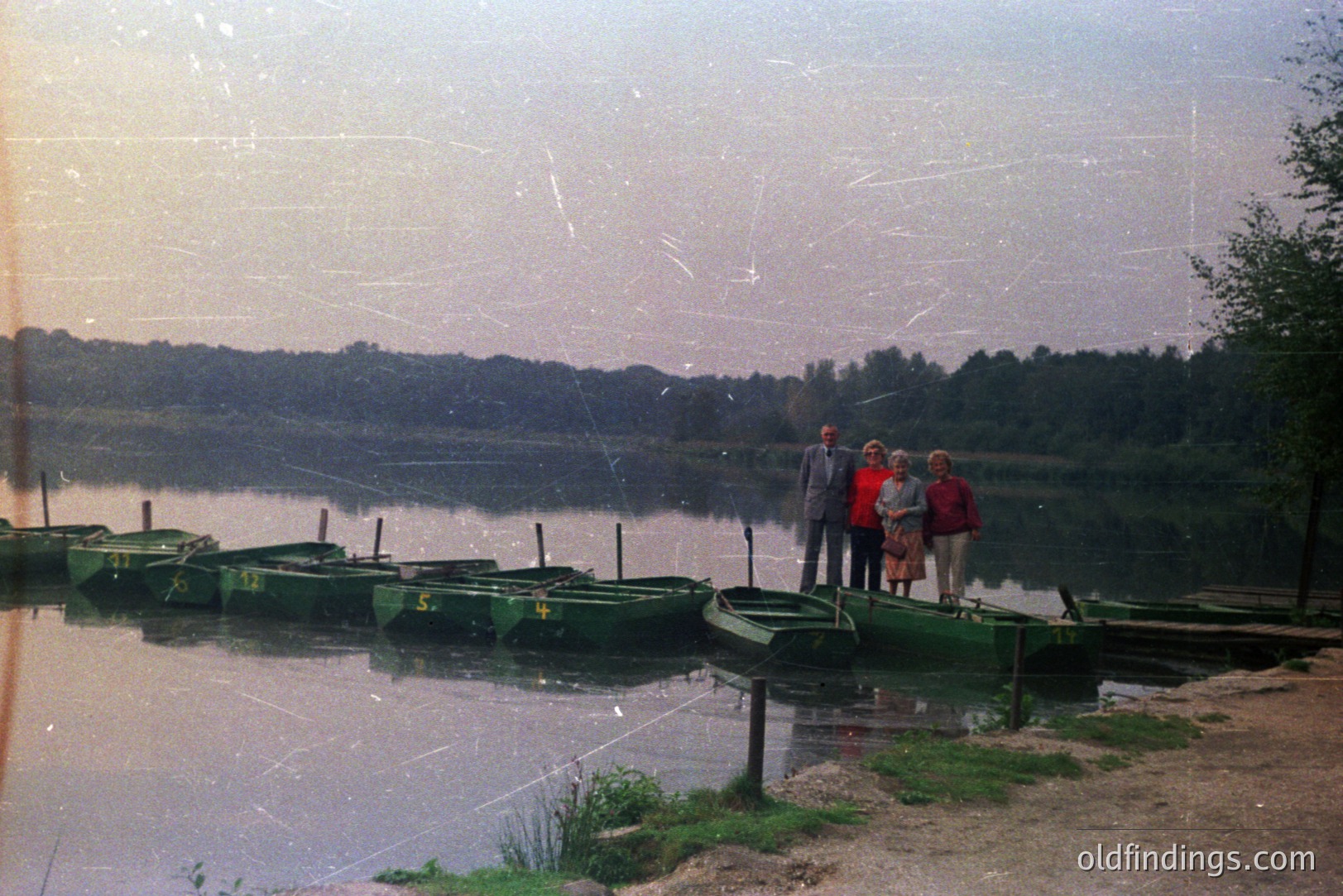A group of three individuals stands along the shore of a lake, alongside a row of green rental boats. The scene evokes a leisure setting, likely a lakeside resort. Appears to be a snapshot from the 1970s based on clothing & photographic style. A tranquil lakeside landscape.