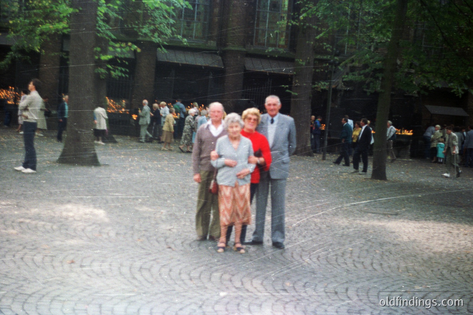 A family portrait: three older individuals stand on a cobblestone plaza, likely a memorial site. Two men flank a woman; all wear classic 1970s-80s attire. Background reveals a large structure with numerous lit candles & onlookers. Appears to be an outdoor gathering.
