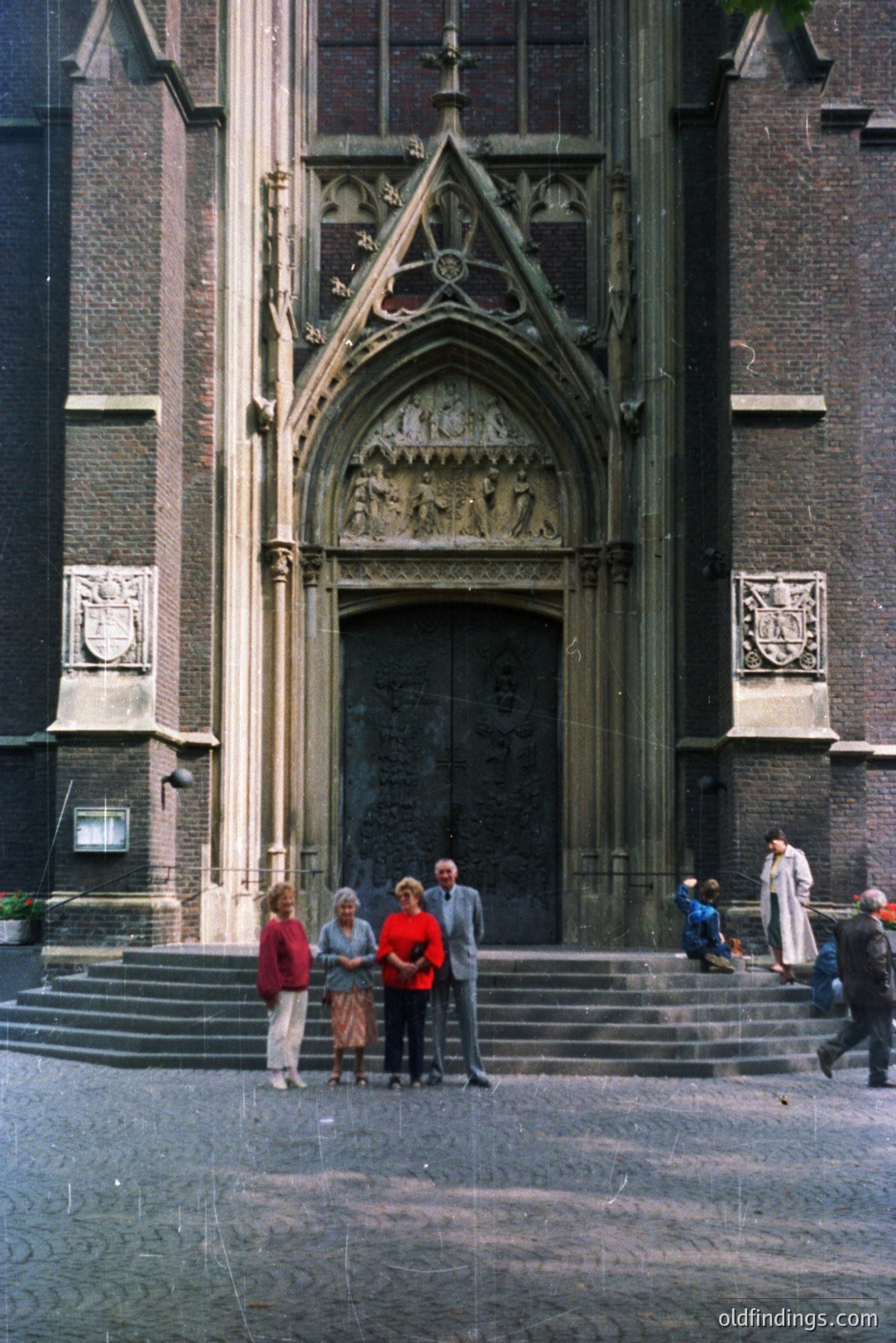 Monumental Gothic architecture features an ornate doorway framed by detailed stone carvings. Three figures stand on the steps, with another person seated and another walking by. Appears to be a church facade. Likely mid-20th century, based on clothing styles.