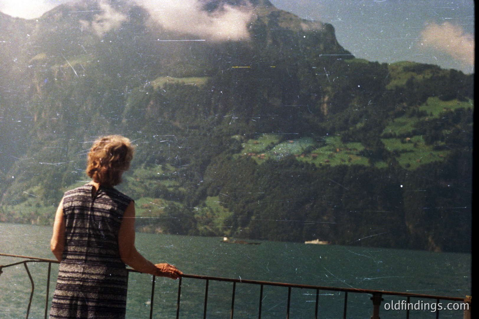 A figure gazes at a fjord landscape from a balcony, possibly in Norway. Lush, steep hillsides descend to a body of water, dotted with small boats. The image exhibits characteristics of mid-century color film, including visible grain and light leaks. A dark, patterned dress is visible on the figure.
