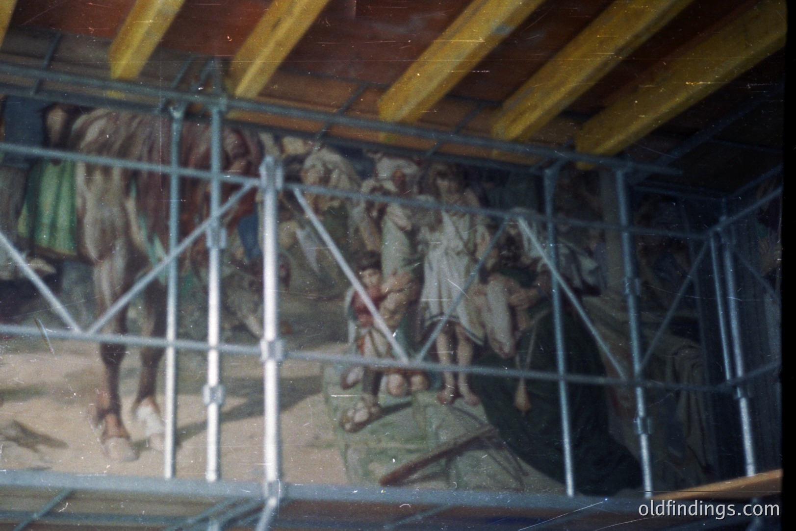 Framed mural depicts figures, potentially biblical or historical, against a landscape with trees and possibly a body of water. A metal scaffolding obstructs part of the view. Likely a restoration or documentation photograph. Colors are muted, suggesting age or film type.
