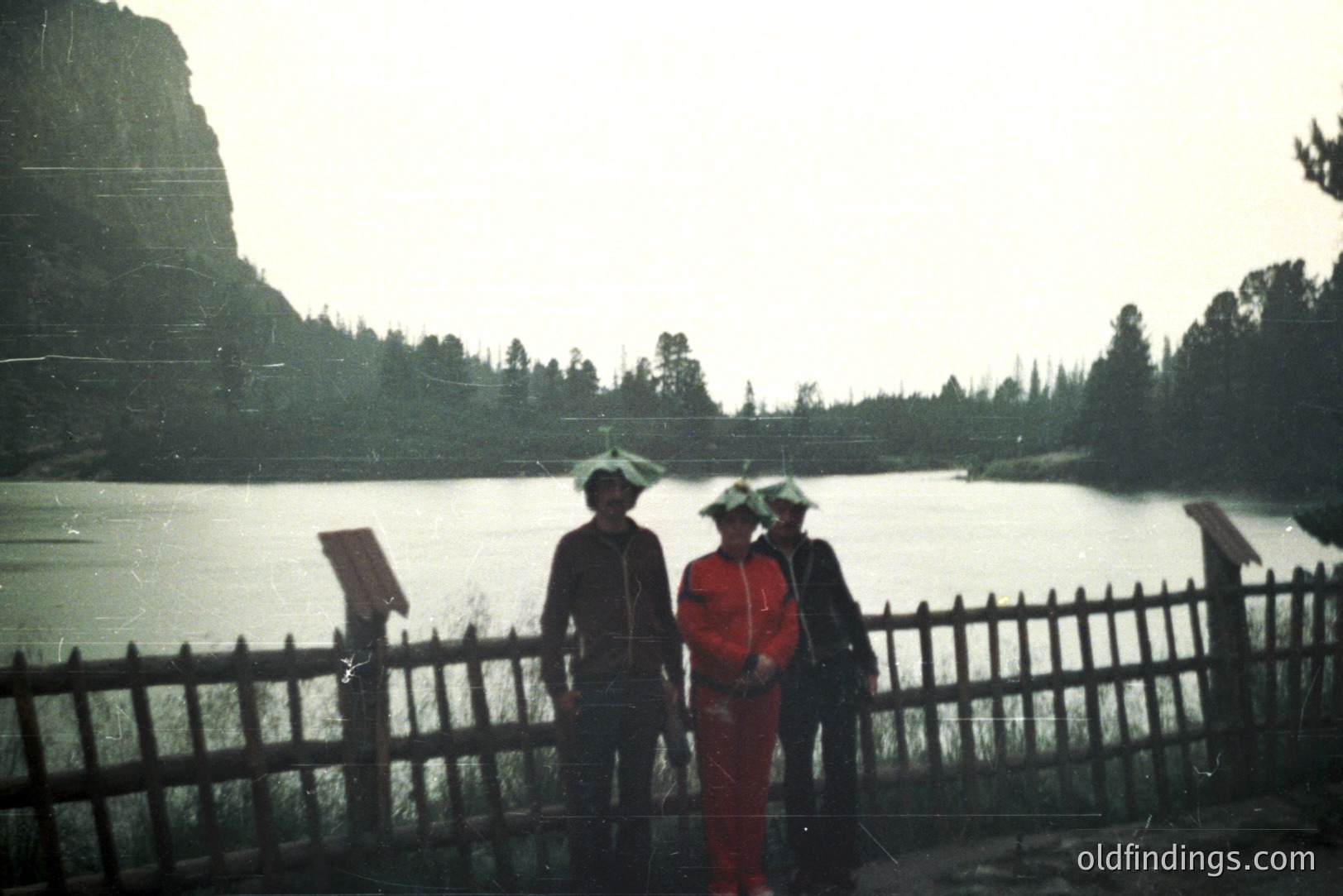 Three figures stand behind a weathered wooden fence overlooking a still, overcast lake backed by a forested hillside. All three wear protective head coverings. The muted color palette suggests a vintage film photograph, possibly 1970s. The scene evokes a remote, possibly industrial, location.
