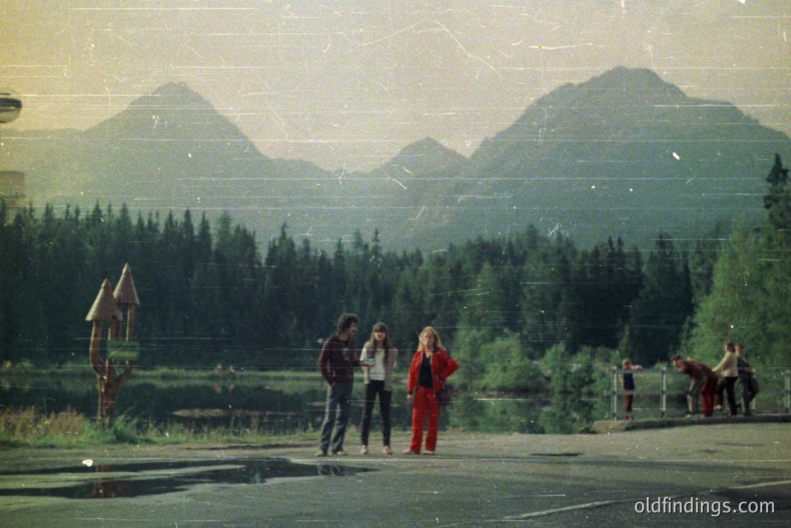 A group of young people stand on a paved road, framed by a dense evergreen forest and towering mountains. The composition suggests a roadside stop, potentially in a mountainous region. Clothing styles, particularly the red snowsuit, hint at the 1970s. Likely a travel or leisure photograph.