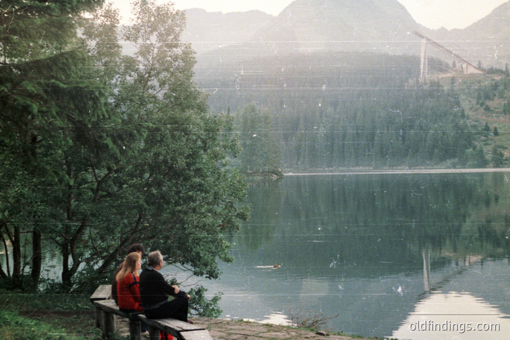 A couple sits on a bench overlooking a calm lake, likely in the Balkan region. Dense coniferous forest lines the shoreline, with a bridge visible across the water. The photo's warm tones suggest the 1970s or 80s, possibly shot on film. A serene lakeside landscape.