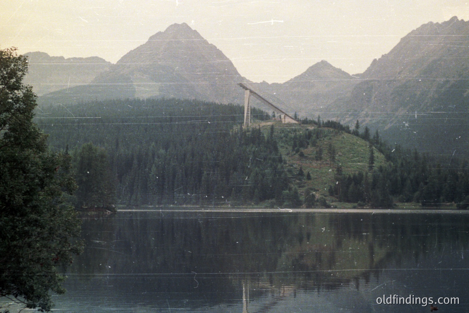 Still lake reflects a towering ski jump structure built on a steep, forested hillside. Dramatic peaks rise in the background, indicating an alpine setting. Likely a promotional or tourism image, possibly from the 1970s or 80s. Appears to be Bulgaria, near Varna.