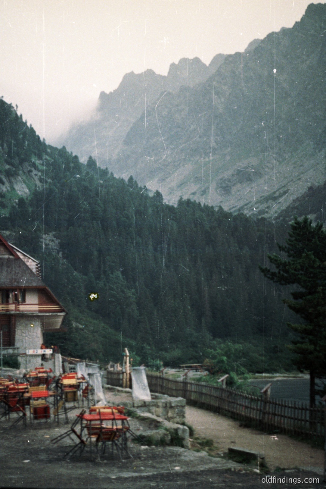 A quaint chalet terrace overlooks a dense forest and steep, rocky mountains shrouded in mist. Rain falls visibly. Red chairs & tables sit empty, with linens draped over some. Likely a Swiss alpine resort scene. Appears to be a vintage tourism image.