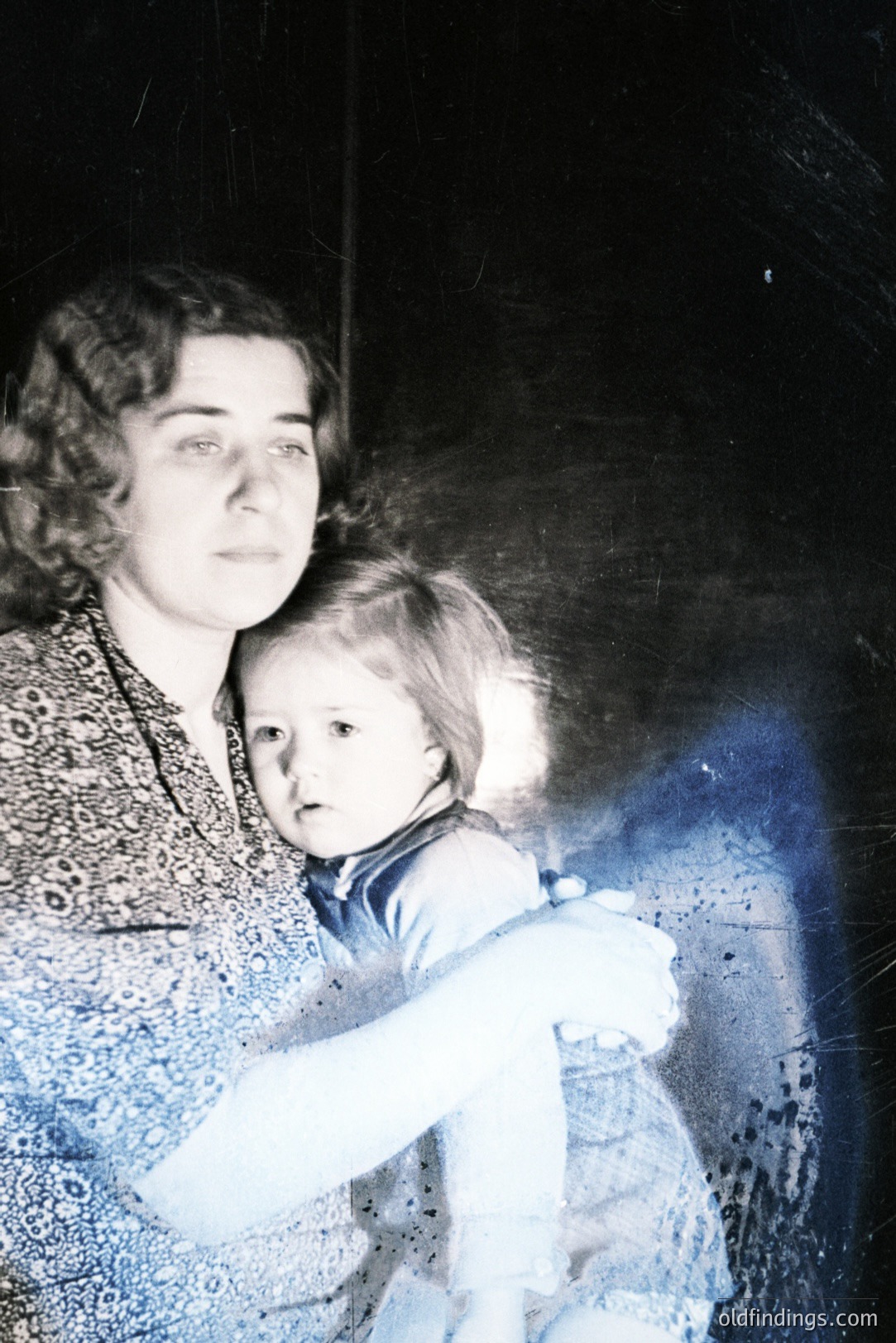 A woman holds a young child, likely a toddler, in what appears to be a home interior. The woman wears a patterned dress, while the child is dressed in blue. The photographic style suggests a 1960s or 1970s family snapshot, exhibiting characteristic color saturation and grain. Image shows signs of aging.
