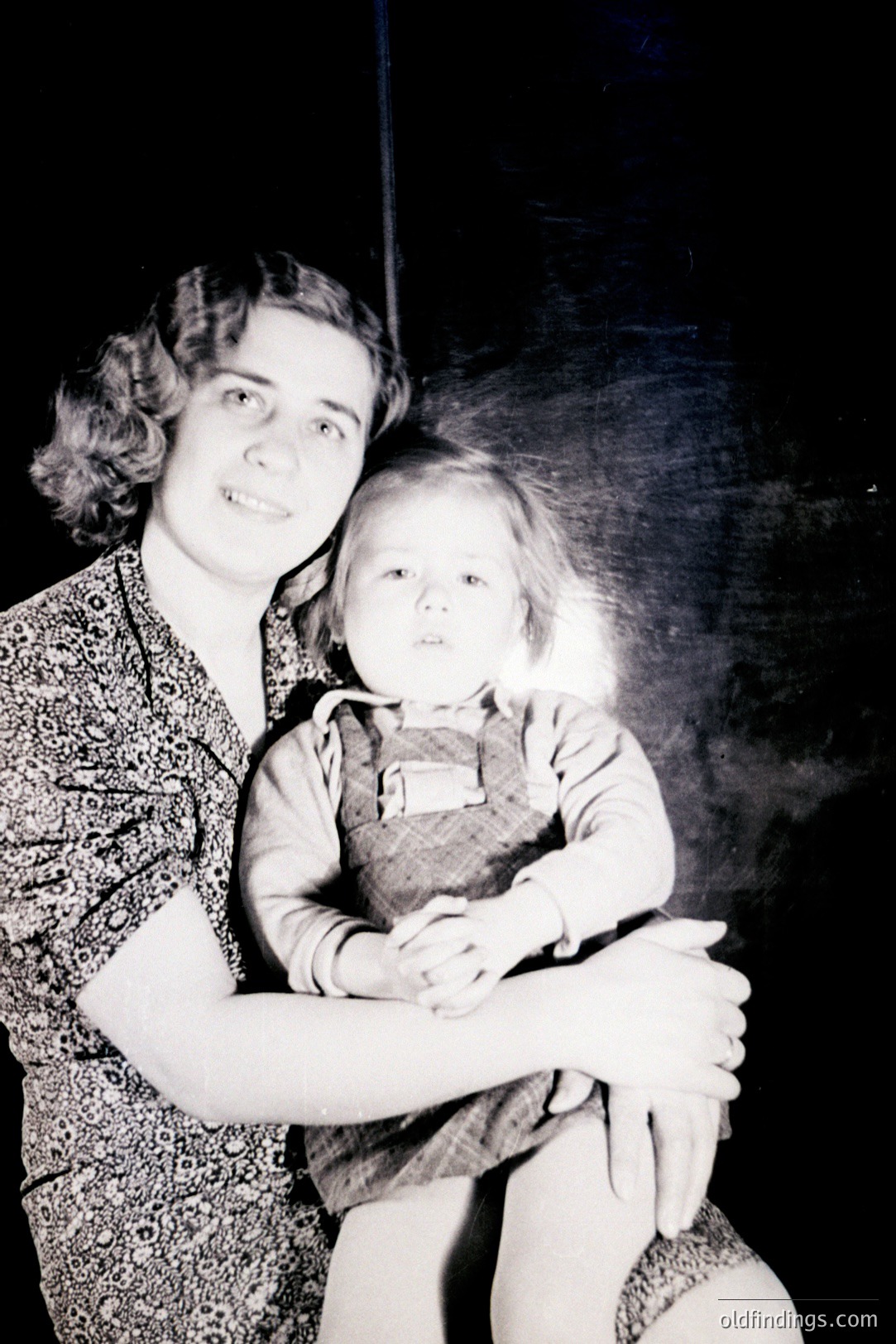 A young woman and child rest on a dark surface, seemingly posing for a studio portrait. The woman wears a patterned dress and has styled hair, while the child is bundled in a sweater. Likely a family snapshot from the 1940s-1950s, displaying a candid moment.