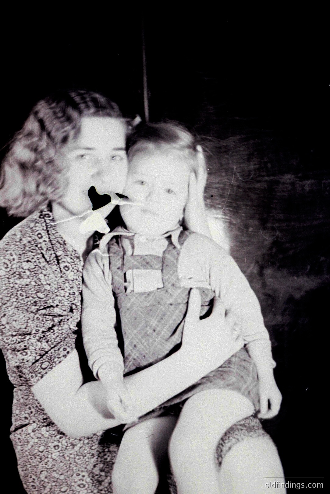 A mother cradles a young child, both with medical tubing visible. The woman wears a patterned dress, while the child is dressed in a checked outfit. Likely a mid-century hospital or clinical setting, evidenced by the backdrop and focused subject.