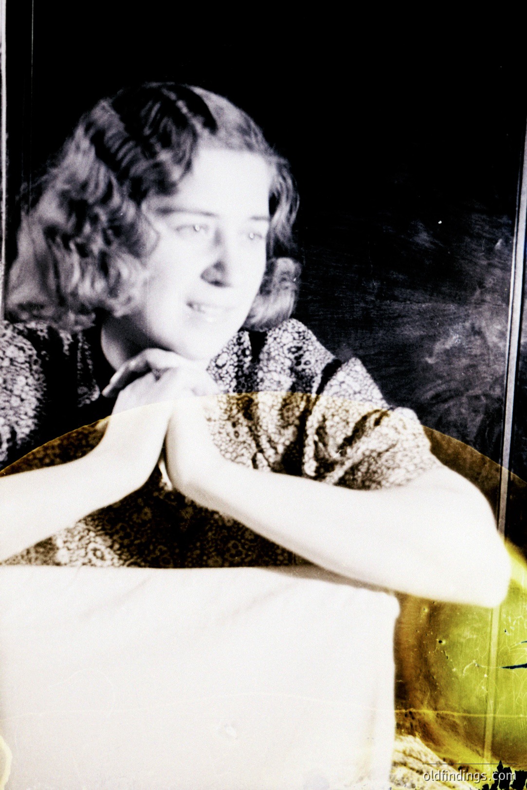 A woman with shoulder-length, wavy dark hair gazes off-camera in this color portrait. She wears a patterned dress, leaning on a table. The backdrop appears to be a dark interior. Likely a 1950s or 60s personal snapshot. Offers reference for mid-century fashion and portraiture.