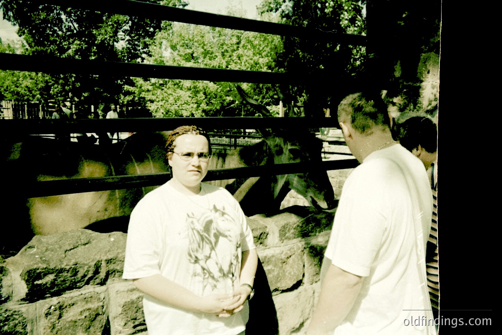 A woman in a graphic tee stands facing a stone wall with a fenced enclosure visible beyond. A man in a white shirt and a partially visible child are to her right. Likely a zoo or wildlife park visit, capturing candid moments. Mid-1990s aesthetic, evident in the film grain & casual attire.