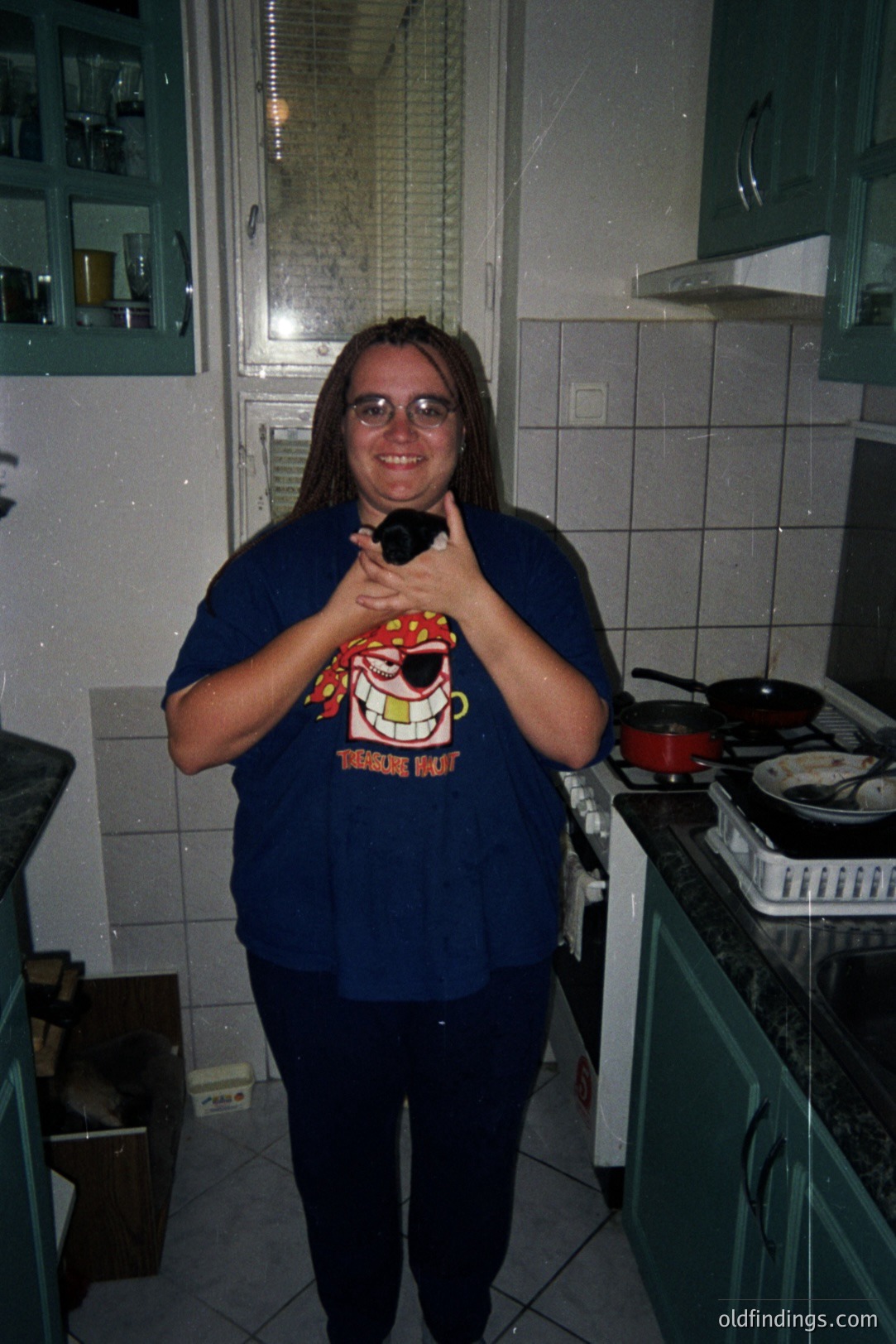 A young man with long hair stands in a kitchen, wearing a dark blue t-shirt featuring a pirate graphic and dark pants. The kitchen showcases green cabinetry, tiled walls, and a stovetop with cookware. Likely from the 1990s, it captures a casual, domestic scene.