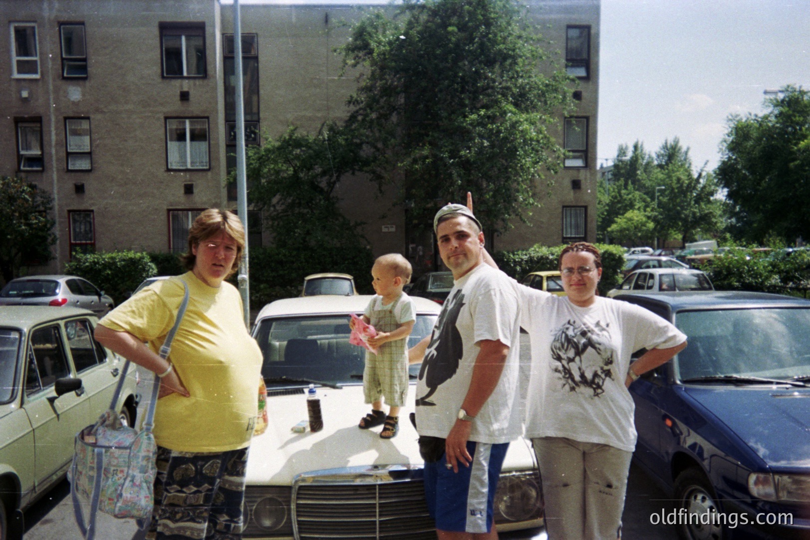A family of four poses near a dark blue car, framed by a large apartment building. The pregnant woman wears a floral skirt and yellow top, while the man sports a graphic tee and shorts. A young child stands between them. Likely 1990s suburban scene.
