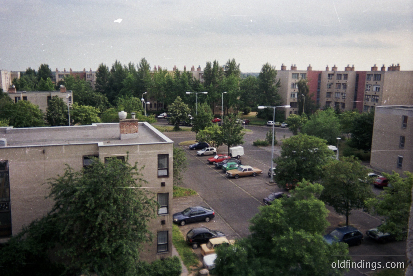 A view of a 1970s-era, high-rise apartment complex in Eastern Europe. Concrete architecture, visible parking lot, and densely planted trees characterize the scene. The location appears to be a planned residential area. A utilitarian aesthetic prevails throughout.