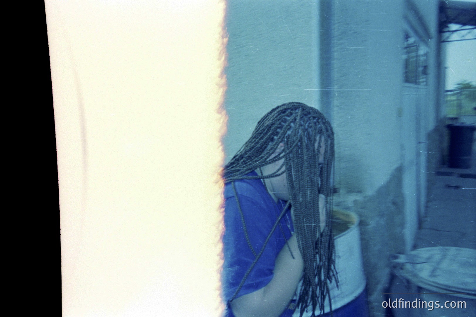 A person, viewed from behind, wears long, dark braided hair and a blue garment, standing near a bucket. The scene appears to be an exterior courtyard or patio, evidenced by a glimpse of foliage and architectural details. Likely a candid snapshot, possibly from the 1990s, given the film quality and clothing style.