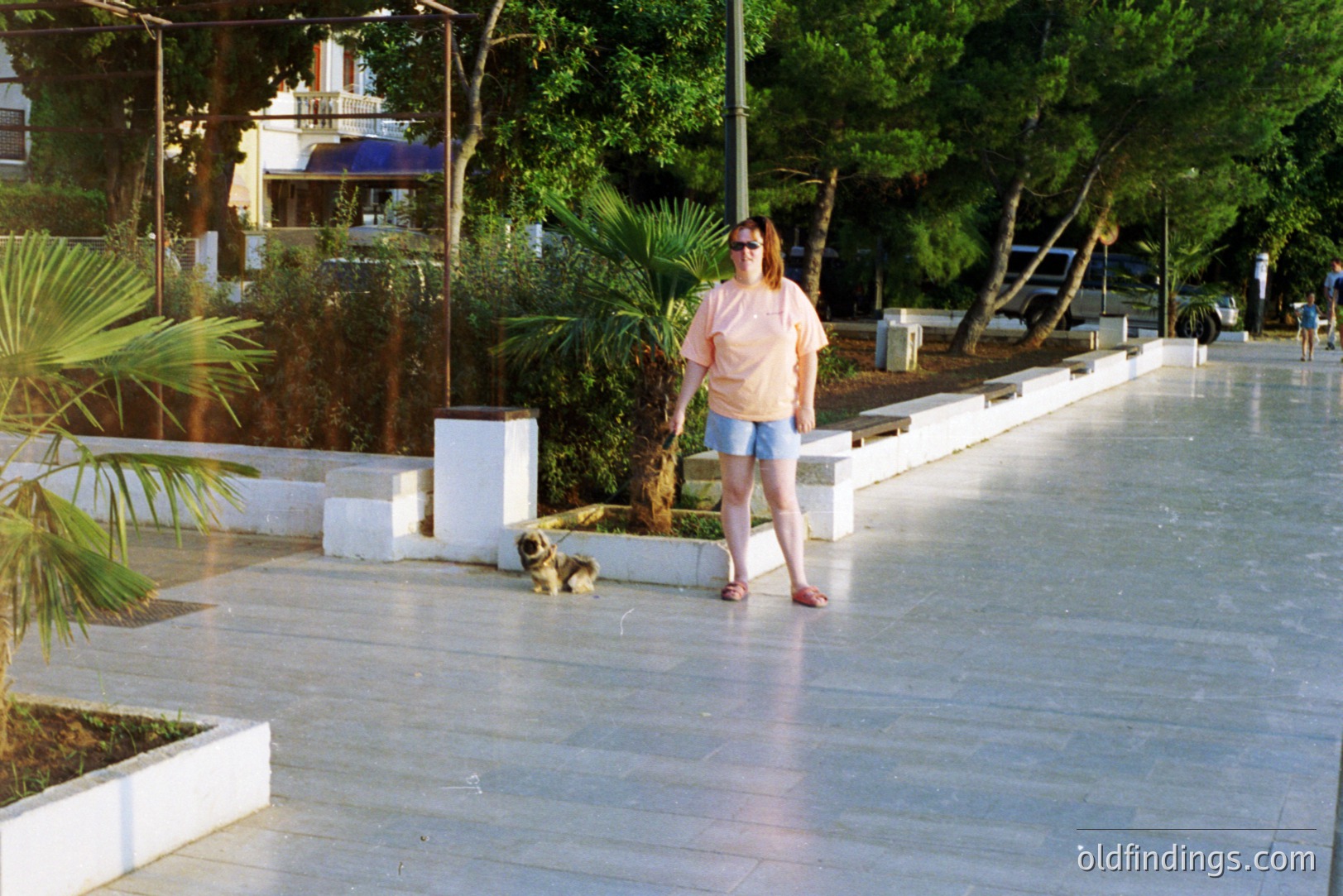 A woman wearing a light peach top & denim shorts stands on a paved seaside promenade. A small, light brown dog sits nearby. Lush vegetation & concrete seating areas define the public space. Appears to be a tourist destination, possibly Southern Europe.