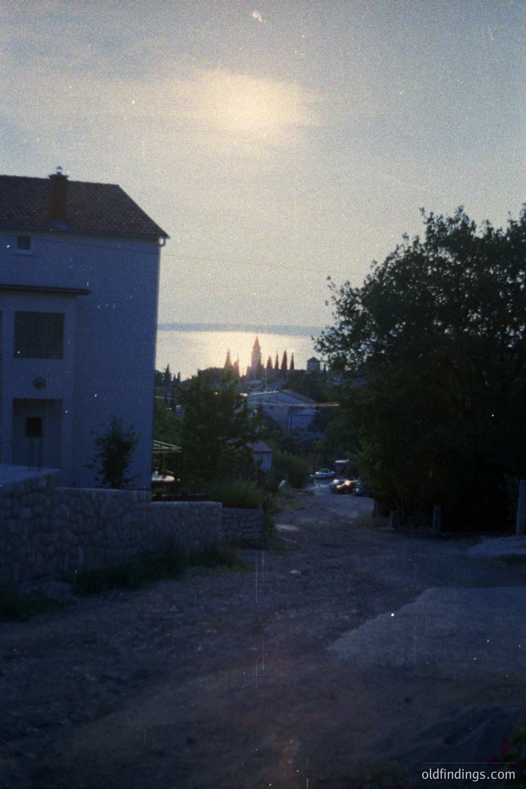 Exterior view of a coastal town at dusk, featuring a two-story white building, stone wall, lush foliage, and a glimpse of a distant waterfront. Appears to be a European locale, possibly the Mediterranean. The scene evokes a tranquil, relaxed atmosphere. Likely 1980s film photography.