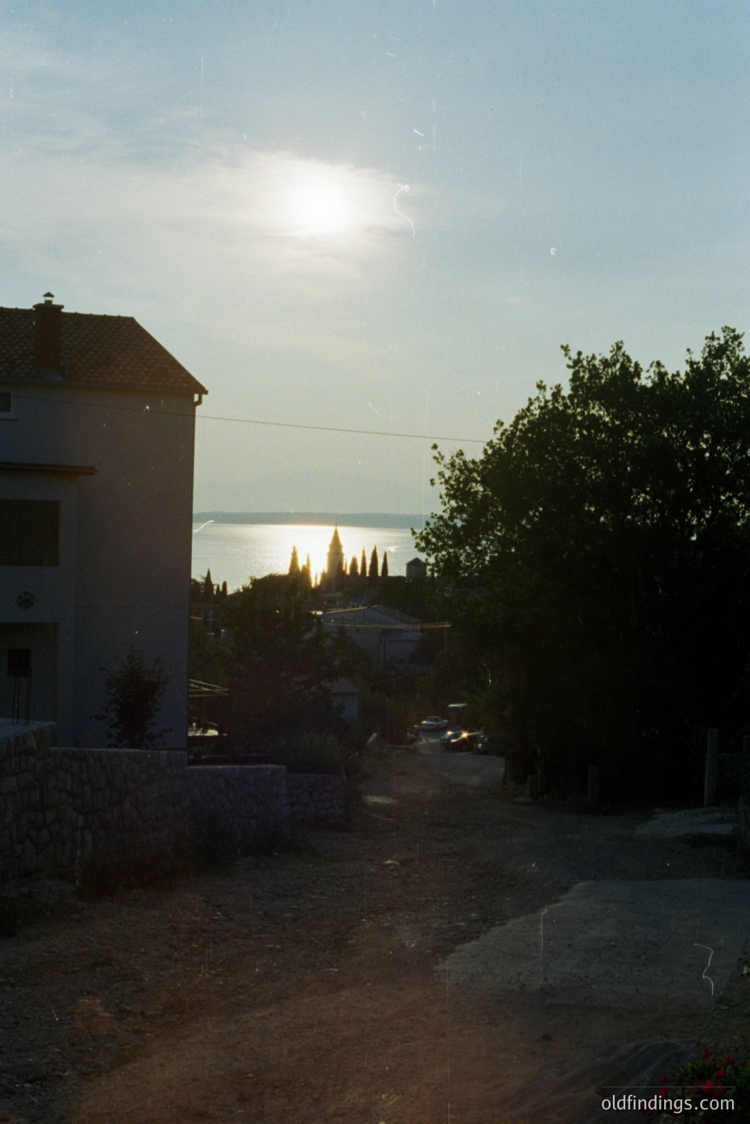 A weathered, two-story building with a tiled roof stands alongside a partially paved road. Beyond, a glimpse of the seaside and a line of trees emerge, framed by a dense green foliage. Likely a coastal European village. The setting sun creates a soft, hazy atmosphere. Likely 1970s or 80s film aesthetic.