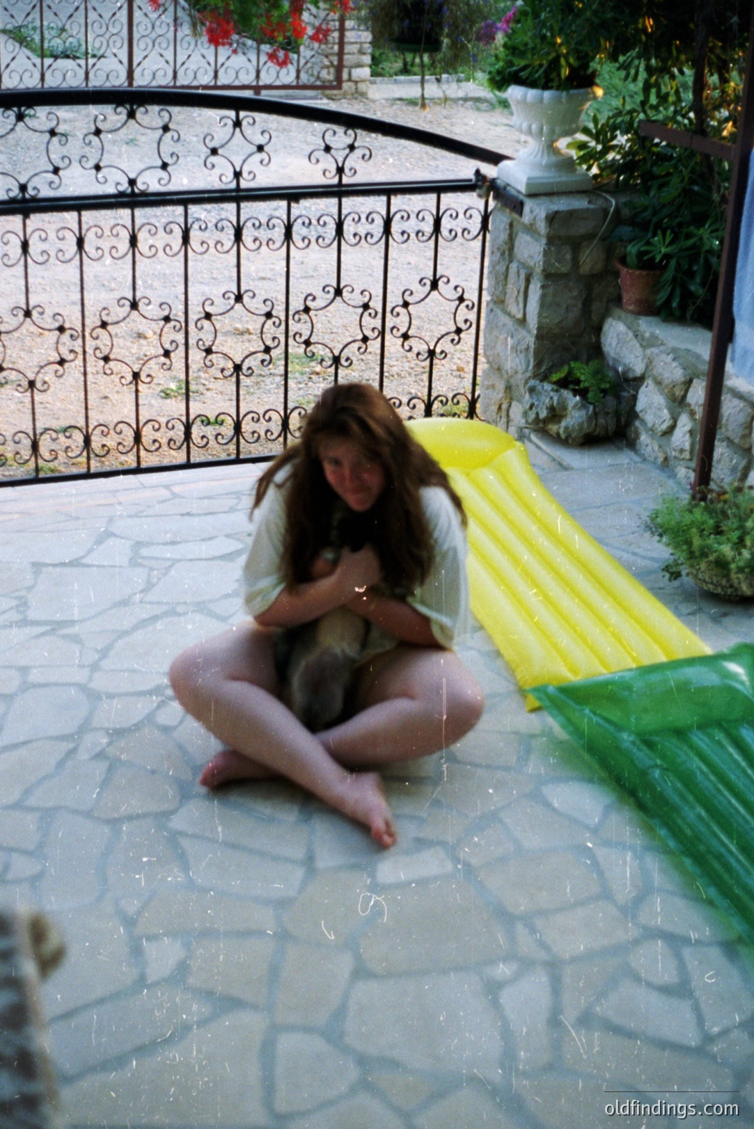 A young woman sits cross-legged on a stone patio, cradling a small dog. Wrought-iron gate & greenery create a courtyard setting. Appears to be a candid moment, potentially late 1990s. Useful for lifestyle or family-themed design.