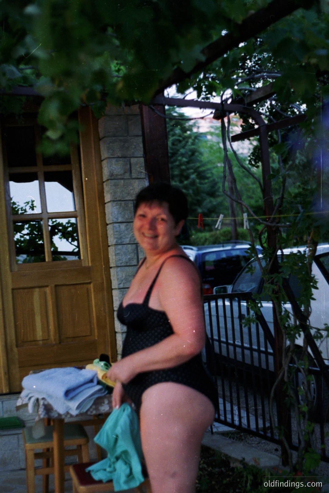 A woman stands outdoors in a black one-piece swimsuit, holding a light blue towel. Stone walls and a wrought-iron gate are visible, hinting at a European setting. Likely a vacation photo from the 1980s or 90s, capturing a casual moment. A glimpse of a car suggests a private residence.