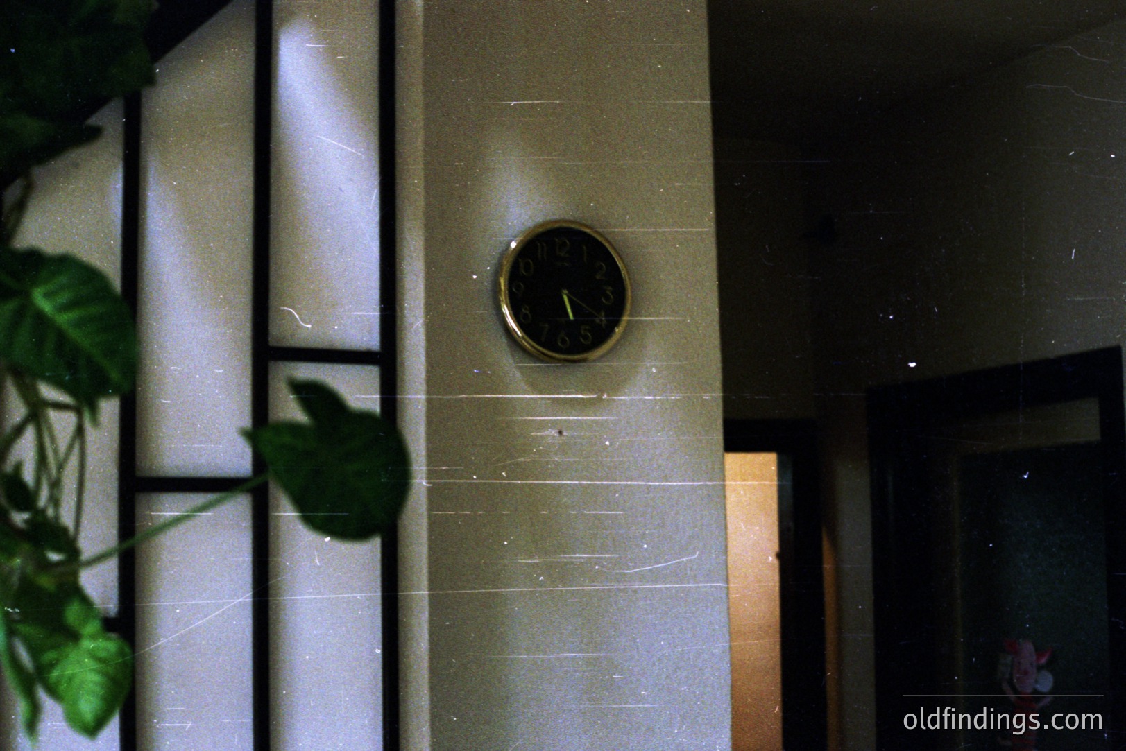 A vintage brass clock is centered on a tiled wall, partially obscured by foliage. The clock face is yellowed with age, showing the time approximately 5:28. The wall appears to be an exterior, likely a courtyard or porch. Possible 1970s aesthetic with film grain.