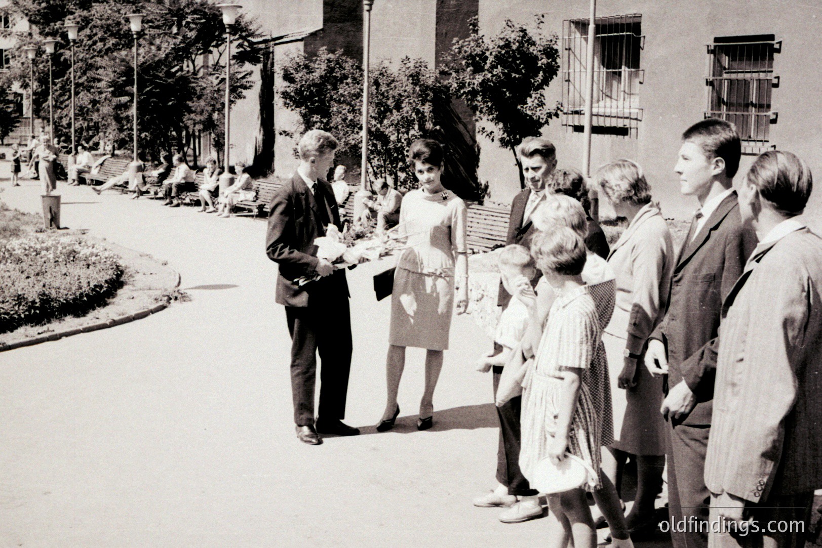 Group of formally dressed young adults and children stand on a paved path lined with greenery and benches. An older couple sits in the background. Likely a school or community event in the 1960s. Appears to be Eastern European, potentially Bulgaria.