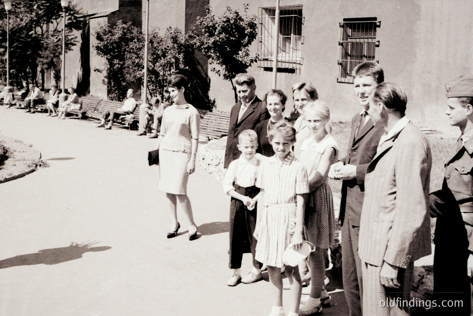 A group portrait of a family and a soldier, posed on a paved street. Architectural details suggest a European setting, possibly a seaside town. The fashion—dresses, suits, and military uniform—indicates a mid-1960s timeframe. A group of seated people are visible in the background.