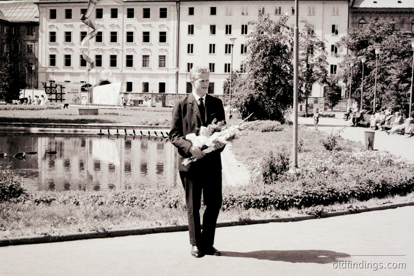 A formally dressed man, holding flowers, stands near a pond reflecting a grand, multi-windowed building. Likely 1960s-70s, possibly Eastern European due to the architecture. People stroll in the background. Suitable for historical context or design.