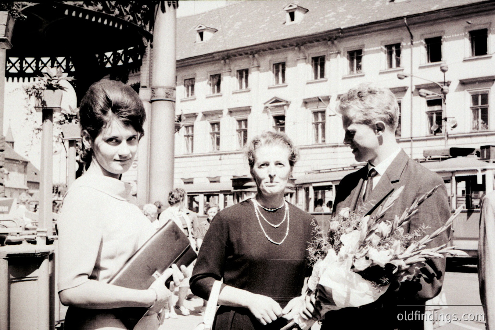 A formal portrait featuring a young woman in a short-sleeved dress, a middle-aged woman in a dark dress with a string of pearls, and a formally dressed man. They appear to be receiving a ceremonial bouquet of flowers, likely an award or recognition. Architectural backdrop suggests a European city square. Appears to be .