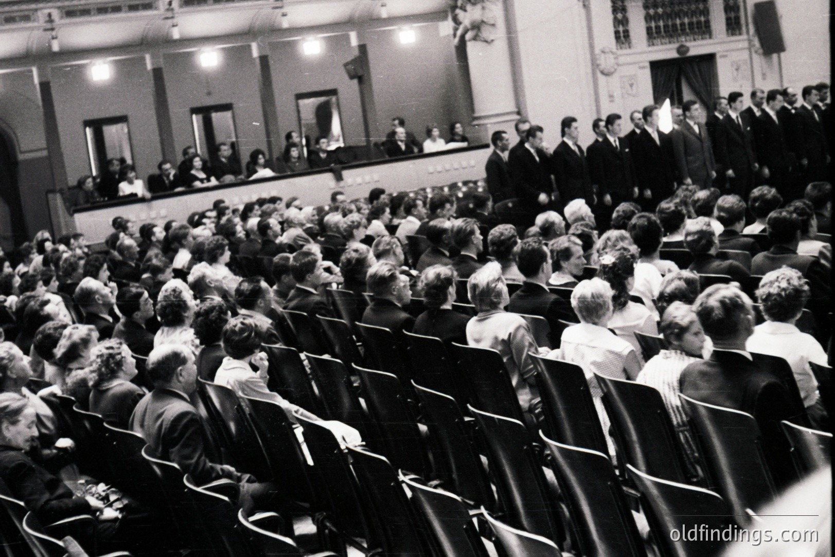 A large audience fills tiered seating, watching a male choral group performing on a raised platform. Formal attire and hairstyles suggest a 1960s or 70s event. Architectural details include ornate ceiling molding. Likely a concert or academic ceremony. Suitable for historical research or design referencing.