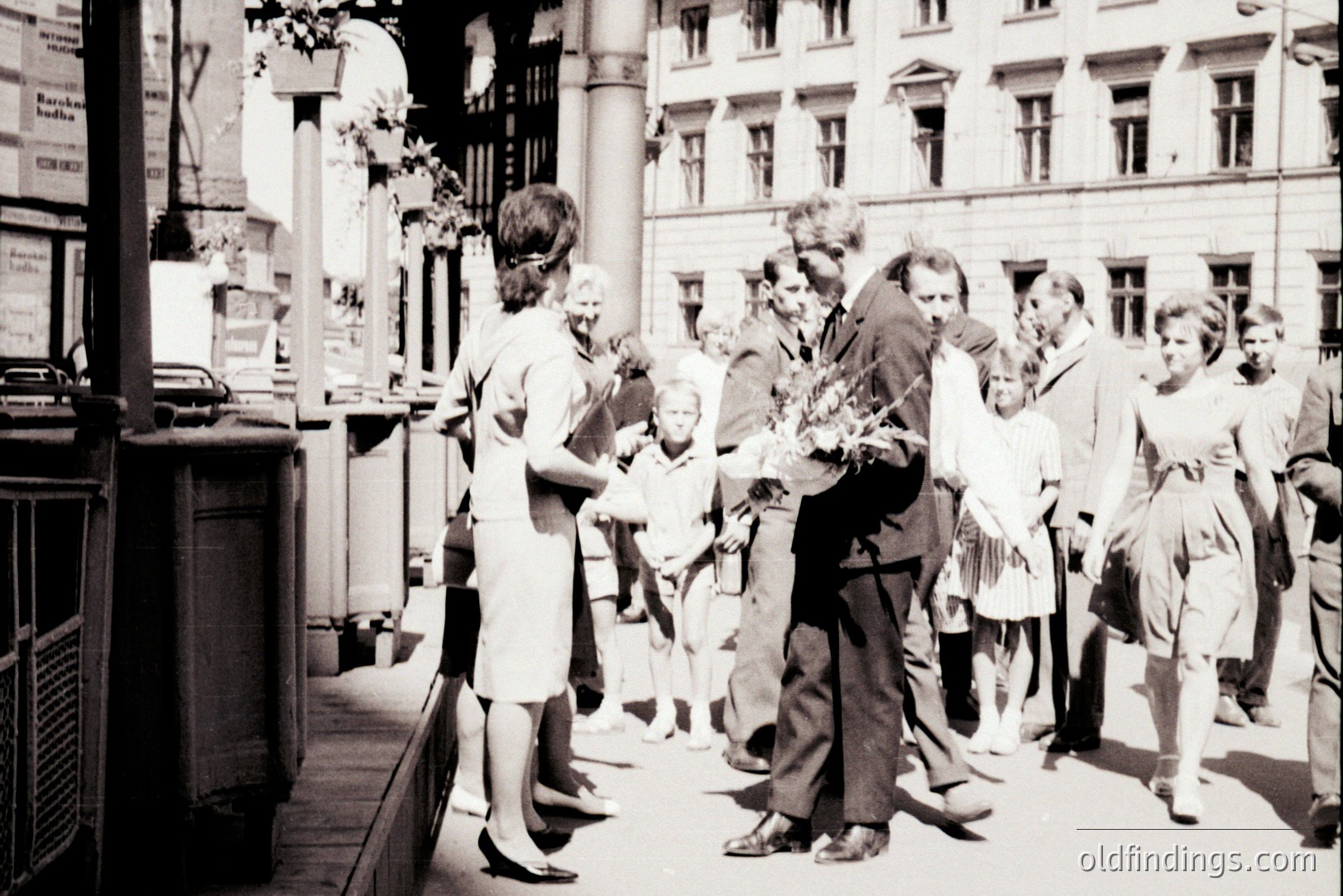 A candid street scene captures a man with flowers interacting with a woman and group of onlookers. Architectural details suggest a European city, possibly Central or Eastern Europe. Appears to be a 1960s or 70s moment, with fashion & vehicle styles reflecting the era. The image has commercial value for stock or design use.