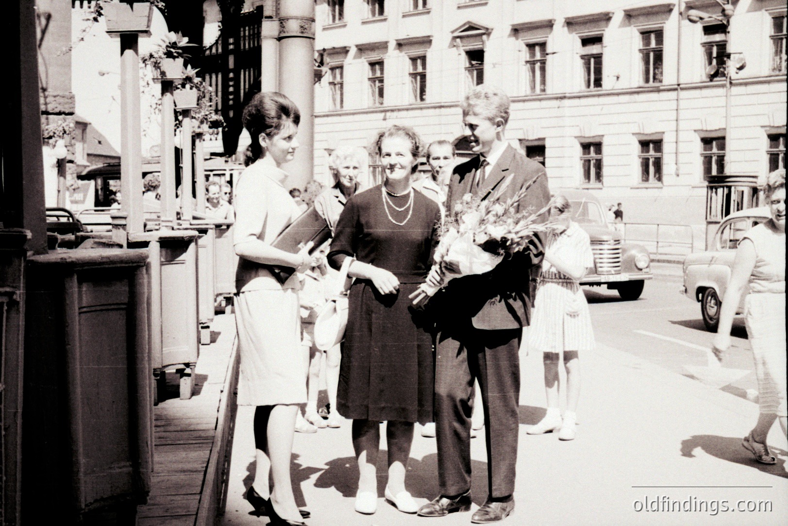 A formally-dressed couple are presented with flowers by a woman in a modest dress. Two young girls in party dresses flank the group on a street lined with classical buildings. Likely a wedding scene, 1960s, Europe. An older automobile is visible.