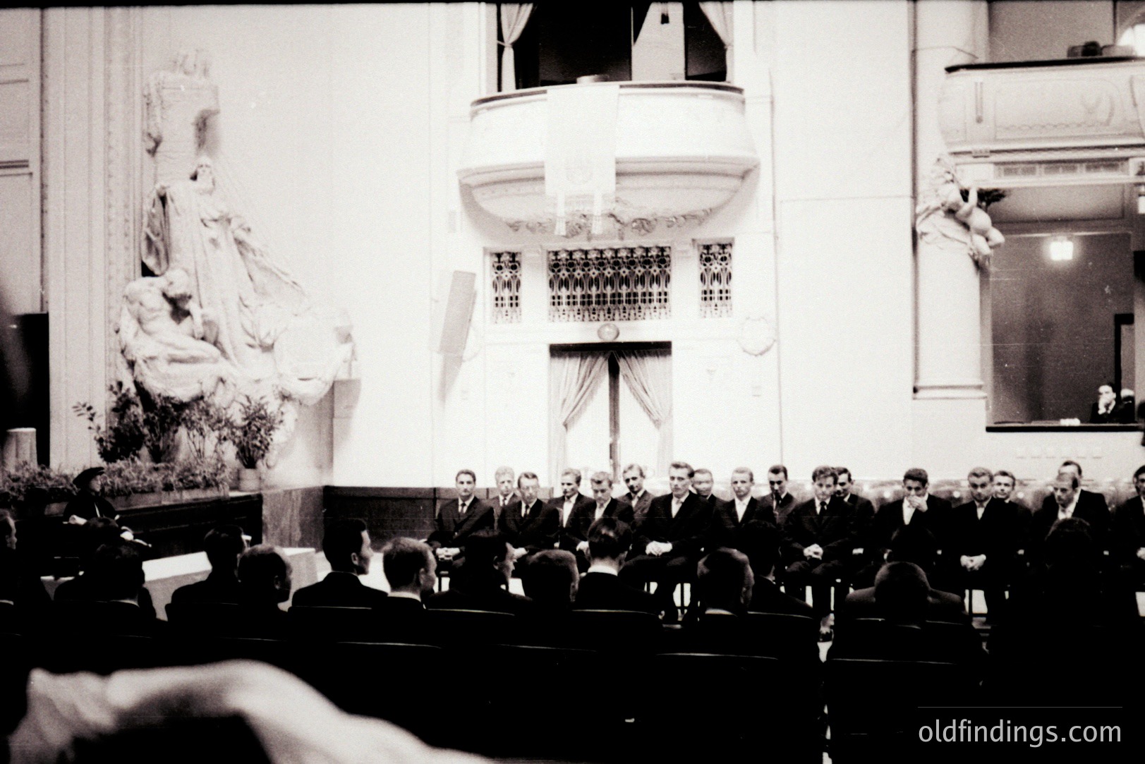 A formal gathering within a grand hall, likely a religious ceremony or official event. A tiered balcony, ornate architectural details, and statues suggest a historic building. Numerous formally dressed men are seated in rows facing a central group. Appears to be 1930s-1950s, based on attire & photographic style. Valuable for documenting societal customs.