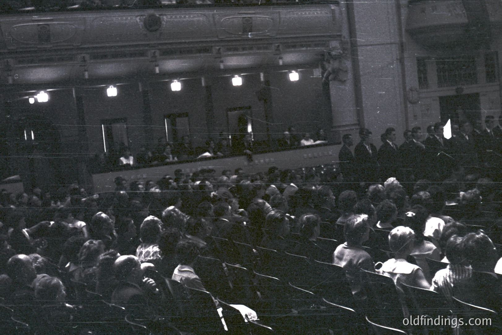 Large audience fills tiered seating within a formal hall, likely a theater or concert venue. Architectural details reveal ornate molding, decorative lighting, and a raised stage area. Appears to be a formal event, with attendees dressed in period attire. Likely 1940s-1960s.