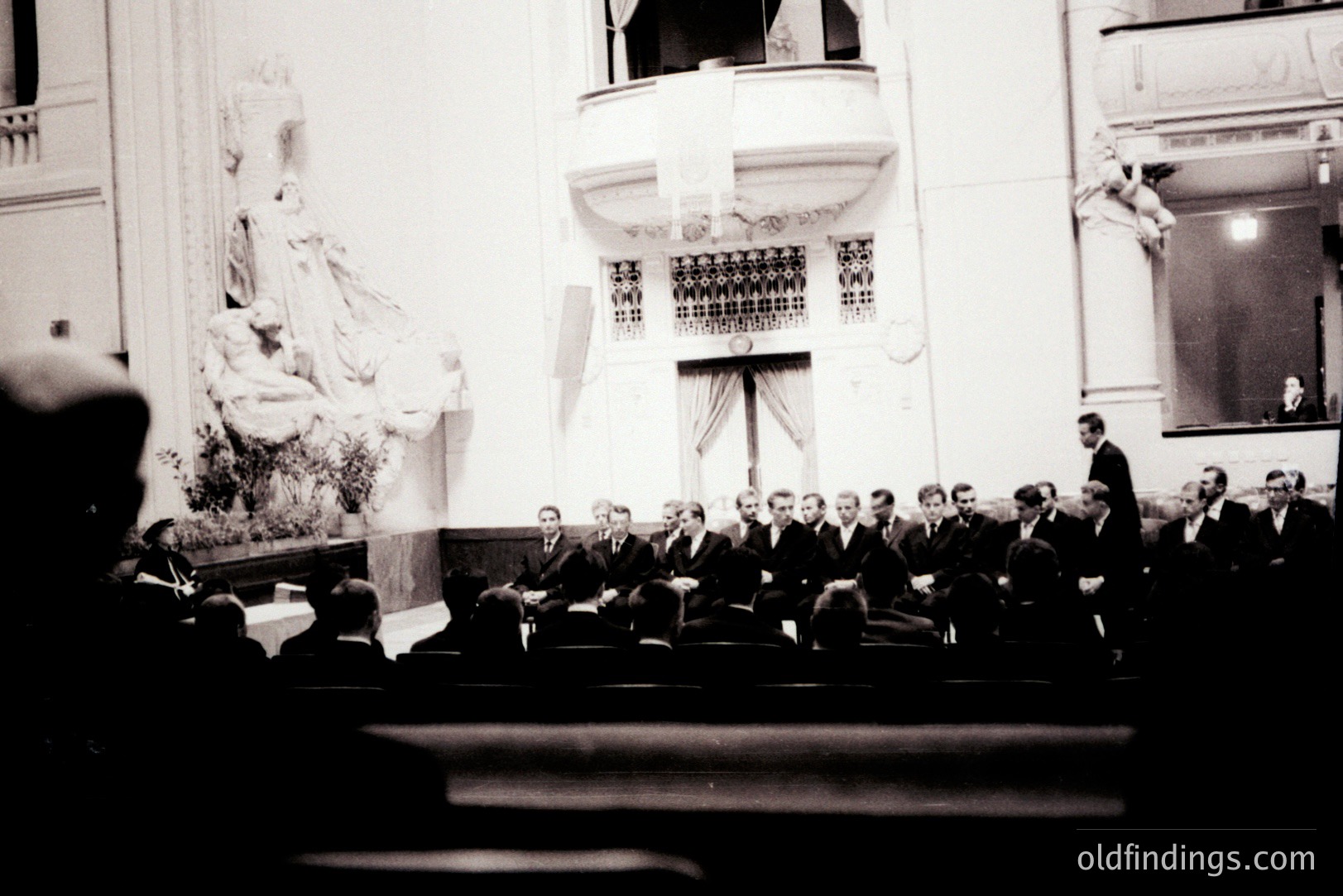 A formal gathering within an ornate building, likely a theater or hall. Numerous men in dark suits are seated in tiered rows. A statue and architectural detailing adorn the background. Appears to be a ceremonial or official event, possibly 1950s-1970s.