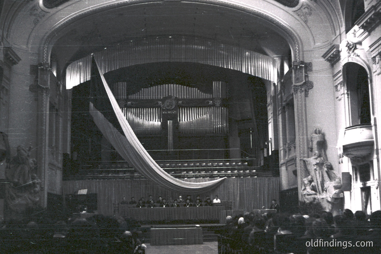 Ornate proscenium stage with a large, draped curtain. Visible audience seated in tiered balconies. Statuary flanks the sides of the scene, typical of an older theater. Likely a performance or formal gathering. The photograph’s style suggests the 1930s or 1940s. A valuable design reference.