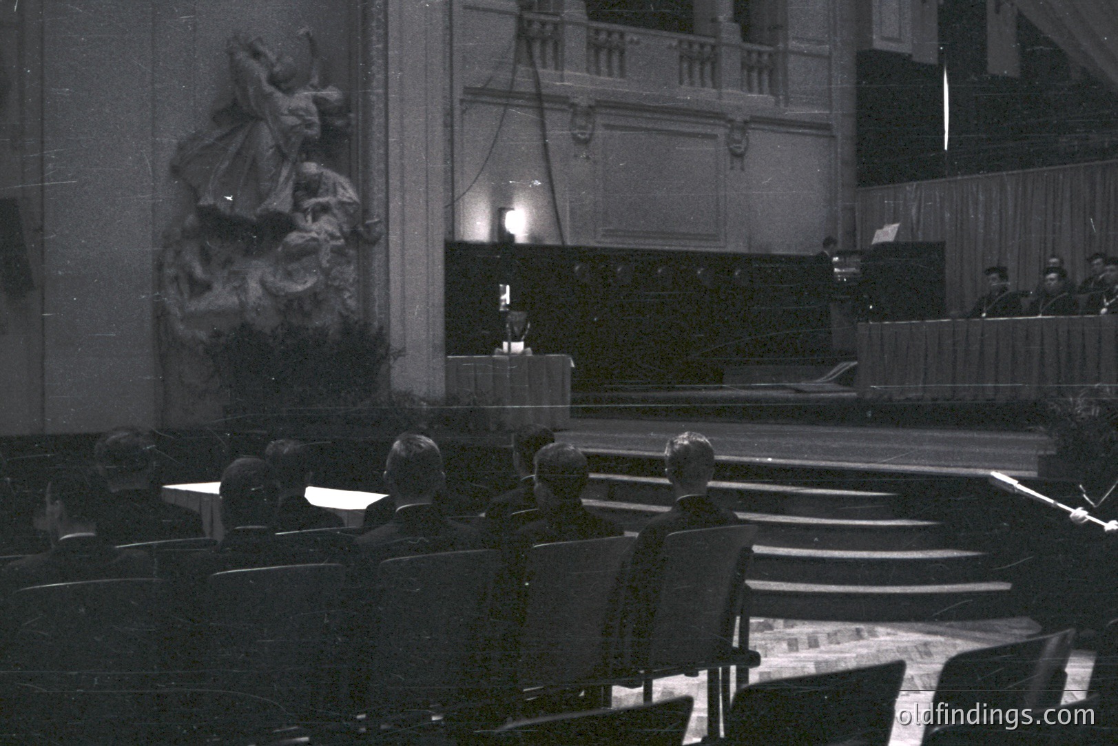 Formal, staged event in a grand hall. Tiered seating leads to a raised platform with a speaker & draped backdrop. A large, ornate sculpture dominates the left foreground. Likely mid-20th century, architectural style suggests European institutional building. Stock potential for design or historical research.