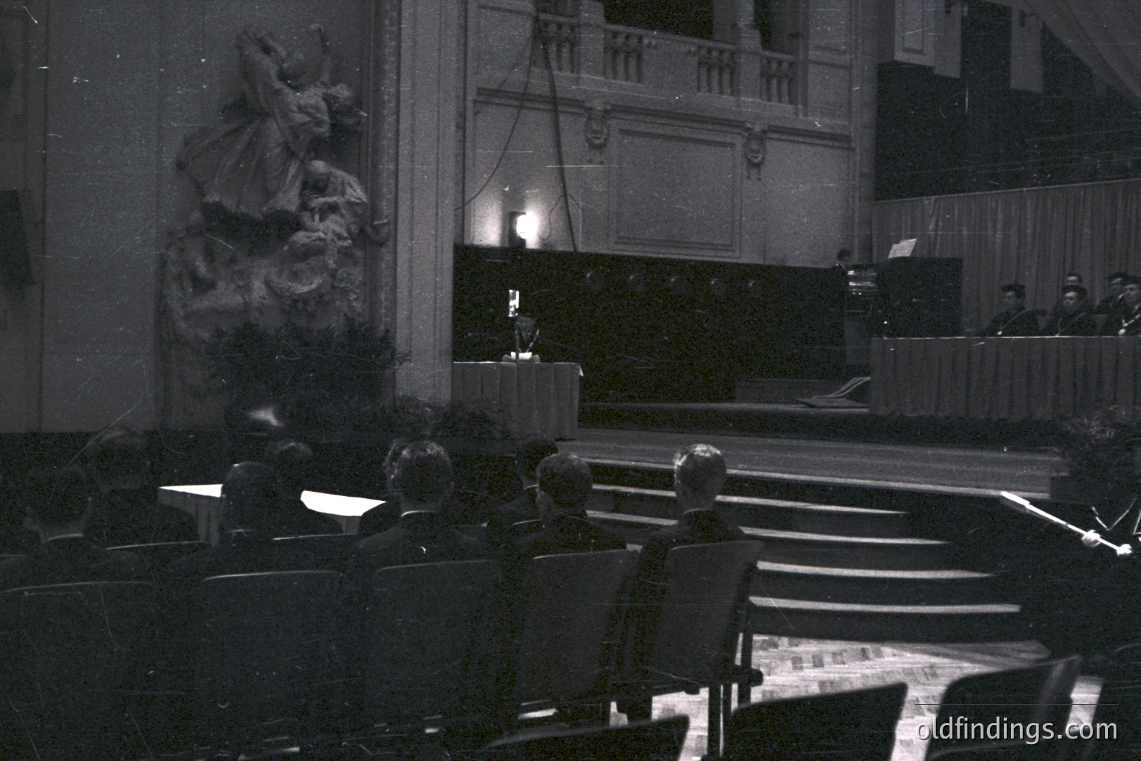 Formal portrait of a group of people observing an elevated stage with a speaker and orchestra. Detailed stone sculpture visible stage left. Architectural elements suggest a grand hall, likely a theater or concert venue. The scene’s monochrome aesthetic points to a 1960s or 70s timeframe.