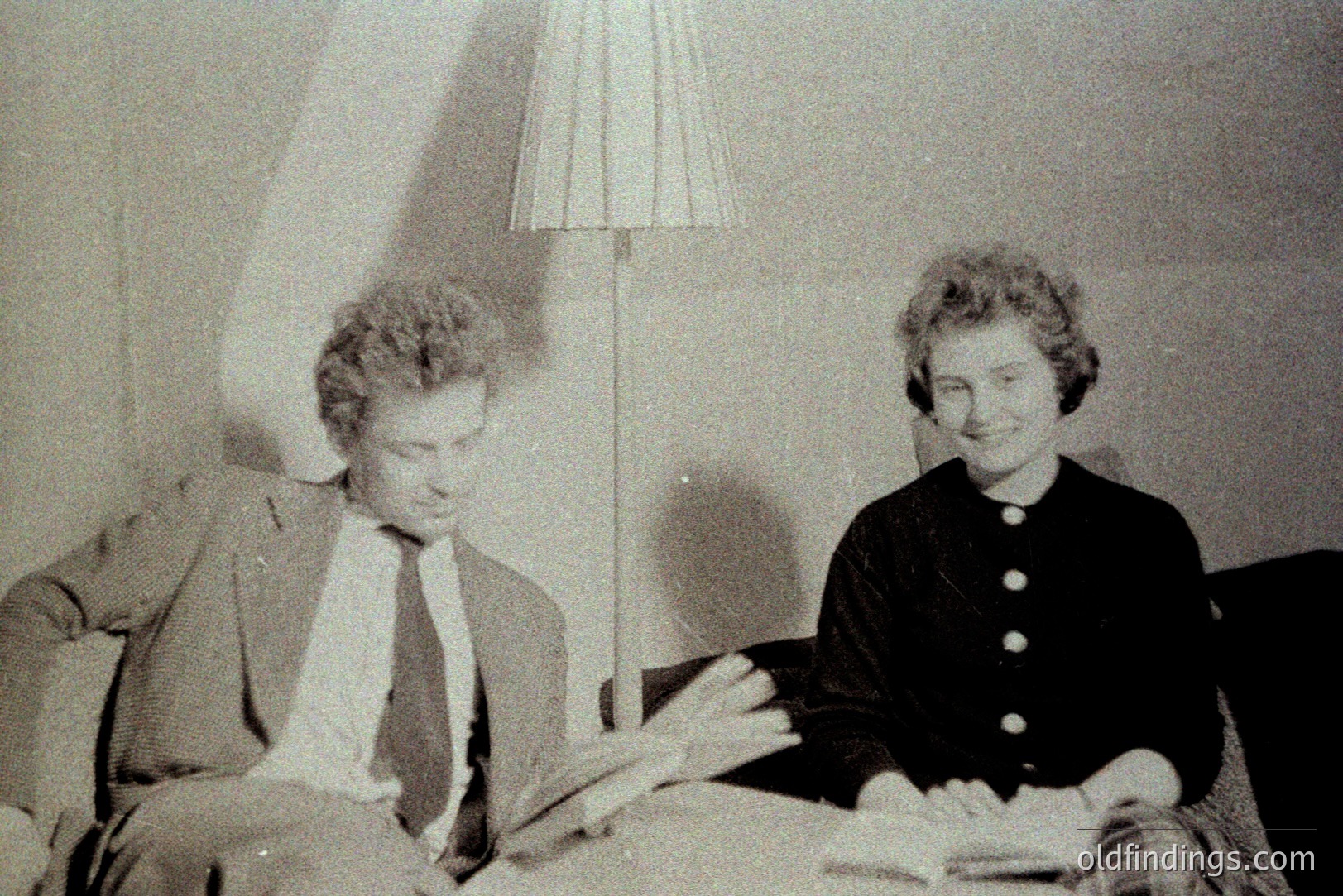 A stylish couple, likely 1960s, pose casually in a domestic setting. The man wears a suit & tie, while the woman's dress features a distinctive high neckline. A floor lamp and apparent books suggest a study or living space. The grain and tonal range indicate potential archival or stock photography origins.