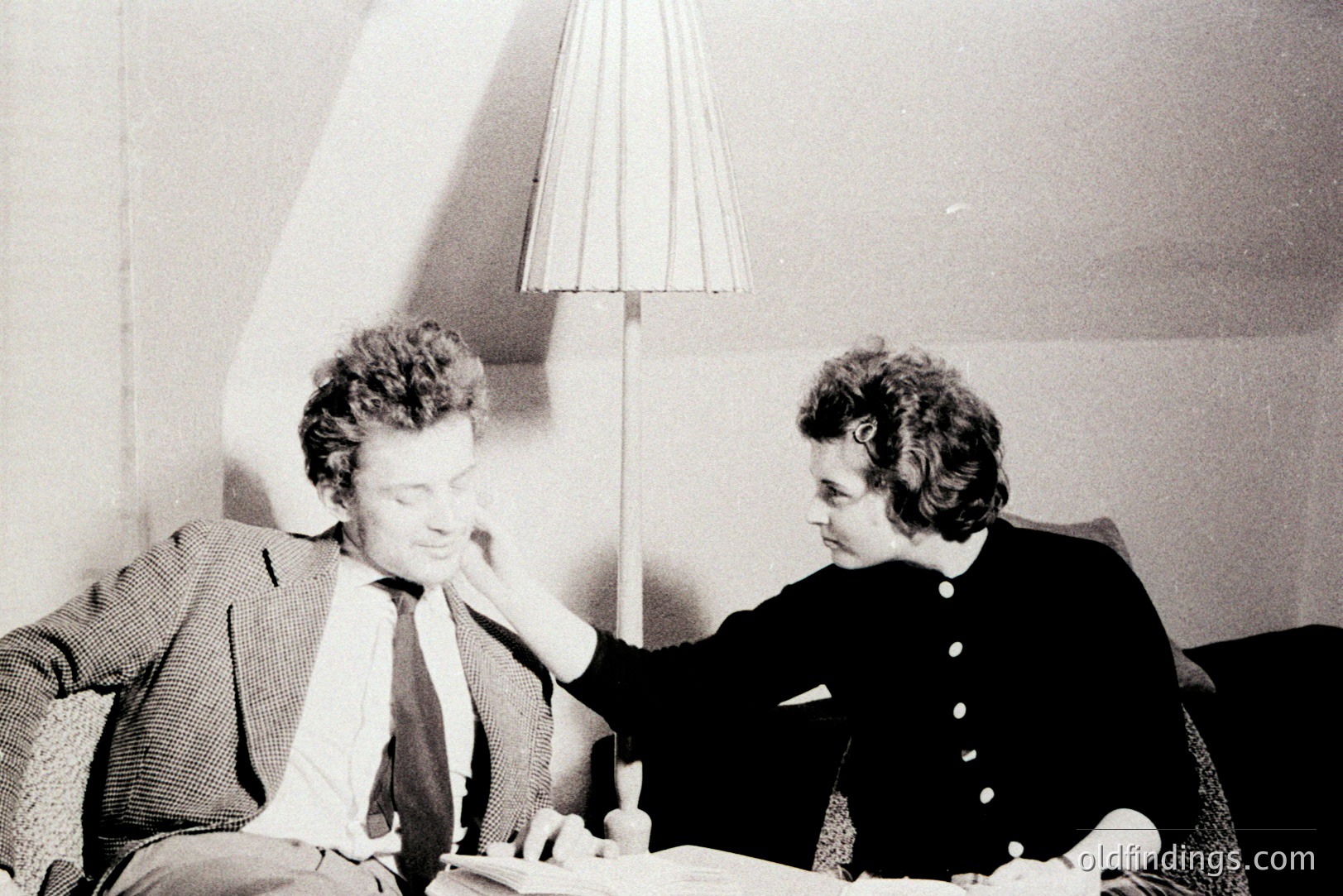 A couple sits closely on upholstered chairs, papers between them. The man, wearing a patterned suit and tie, rests his head on his hand. The woman, in a buttoned dress, leans towards him. A tall floor lamp illuminates the scene. Likely a candid portrait, mid-1960s aesthetic.