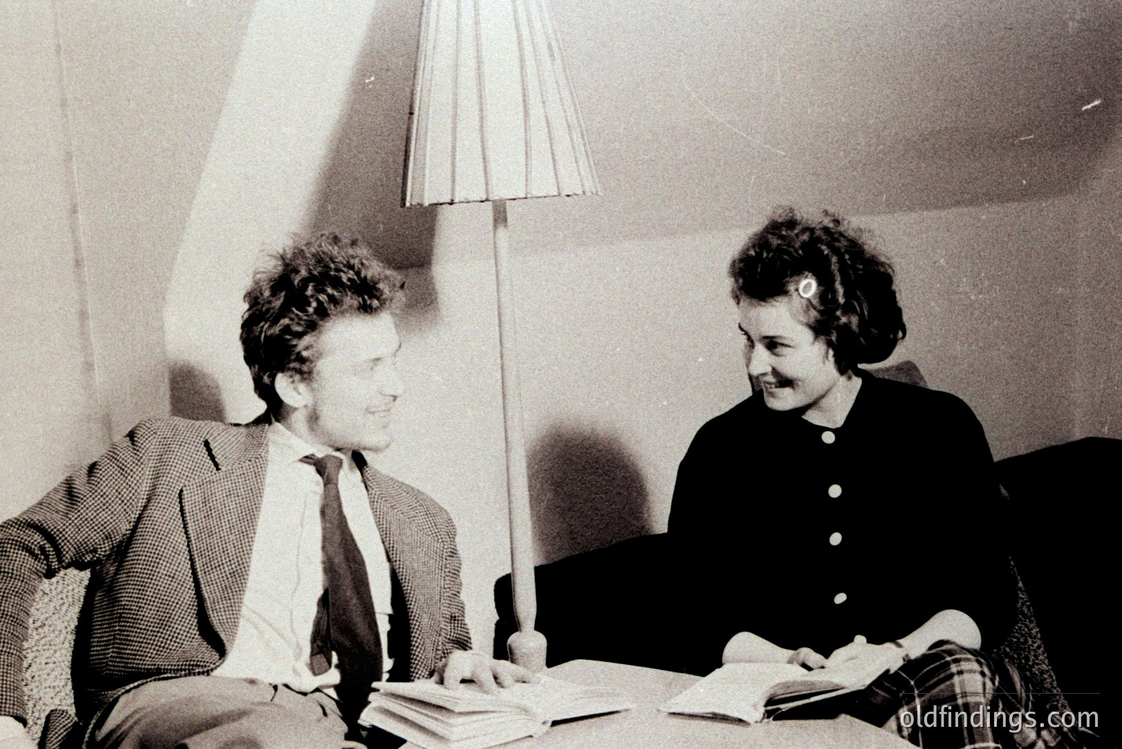 A young couple sits casually in a room, books spread on a table between them. The man wears a patterned jacket and tie, the woman a dark dress. Soft light from a lamp illuminates the scene, suggesting an intimate study or discussion. Likely 1960s, mid-century design aesthetic.
