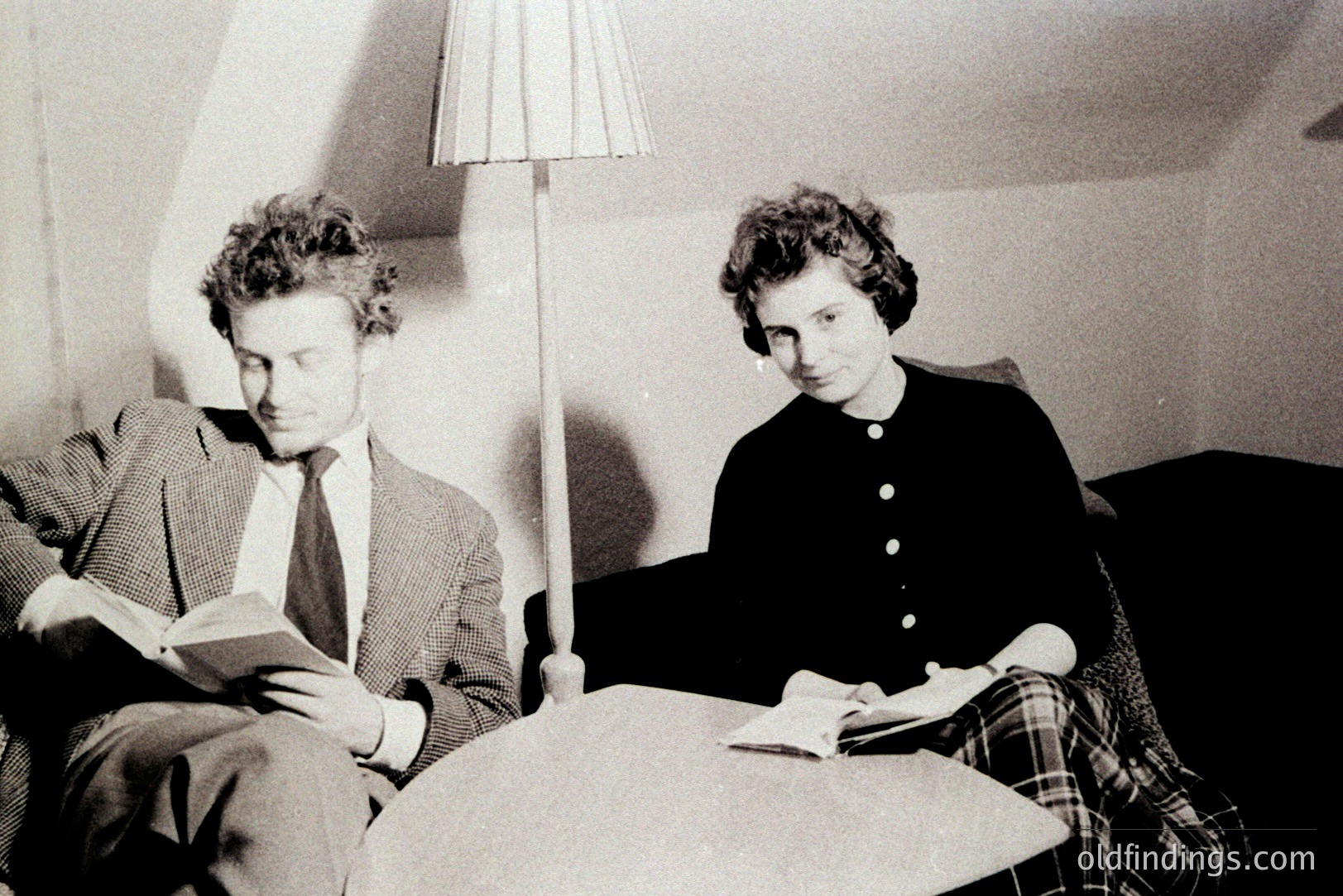 Two figures, a man and a woman, are seated reading books in a warmly lit interior. The man, in a checked blazer and tie, leans back on a sofa, while the woman sits at a small table, also engrossed in a book. Likely 1960s style. A tall lamp illuminates the scene.