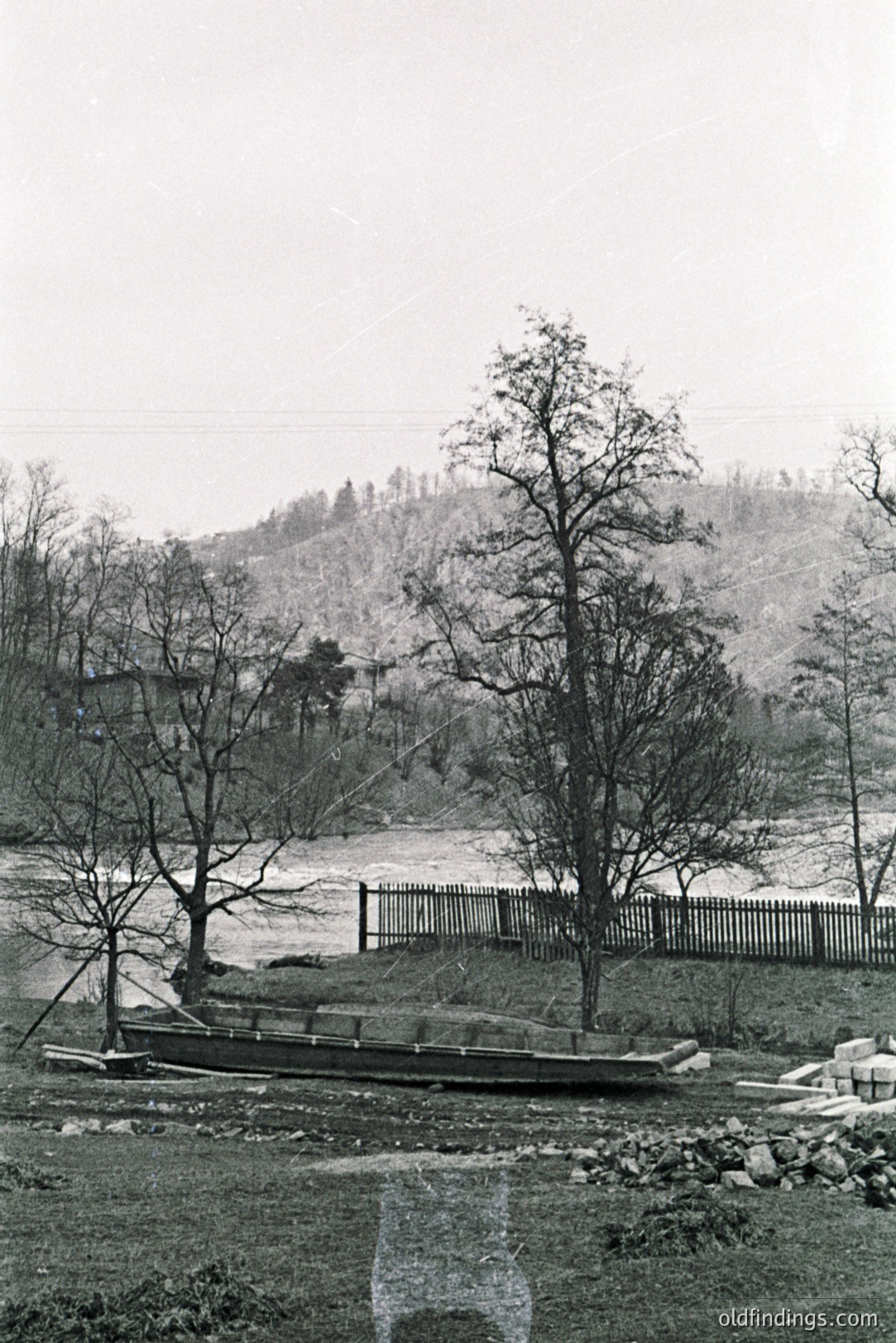 A rustic scene depicts a wooden boat resting on grassy ground near a waterway, framed by bare trees and a weathered wooden fence. Sloping hillside vegetation forms a backdrop. Likely rural location, possibly Eastern Europe. Appears to be an older, possibly archival, photograph.