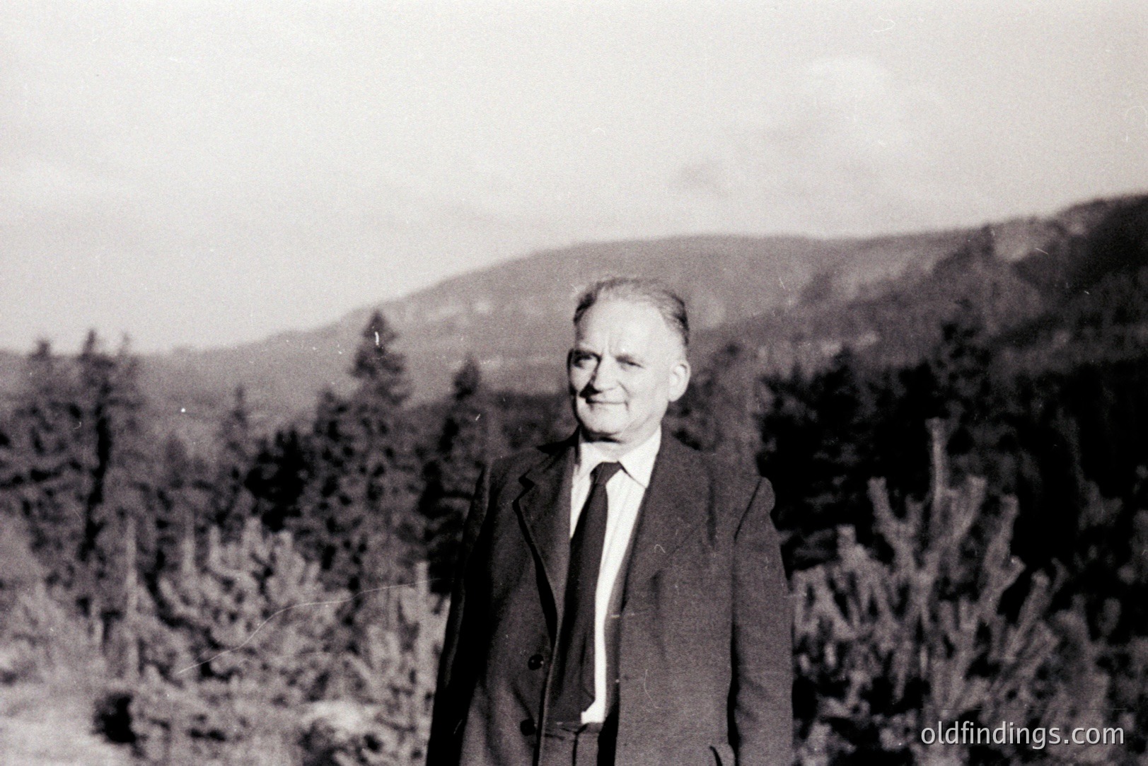 A portrait of a formally dressed man standing amongst evergreen trees, likely in a mountain setting. He wears a dark suit, tie, and overcoat, appearing well-groomed. The background shows a gently sloping hillside and a distant peak, creating depth. Appears to be from the mid-20th century, perhaps 1950s-1970s. A common family photograph style.