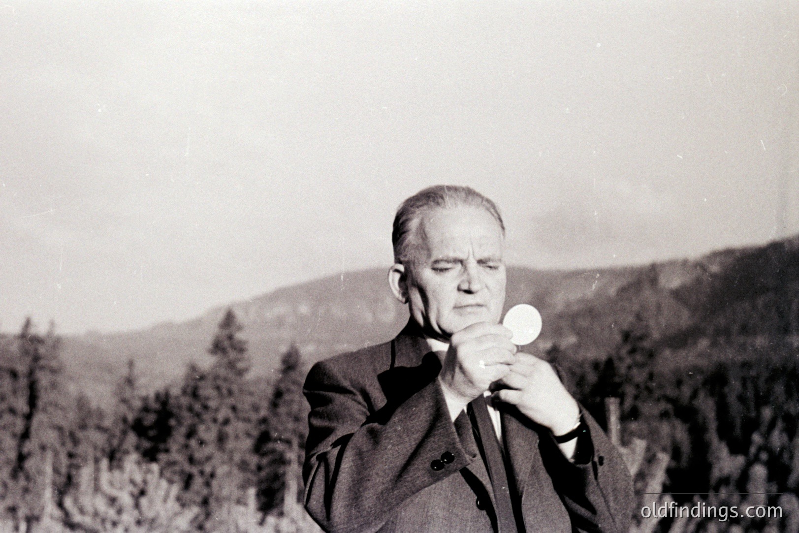 A formally dressed man, likely a priest, holds a host during what appears to be an outdoor religious ceremony. Coniferous trees and a distant, hazy mountain range form the background. The image’s style and tonal range suggest a mid-20th century (1950s-1970s) origin. A poignant, solemn moment captured in black and white.